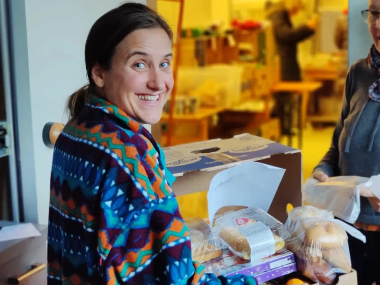 Eine Frau, die lächelt und hinter einem Tisch steht, auf dem Sandwiches und andere Lebensmittel stehen. Im Hintergrund sind Menschen zu sehen.