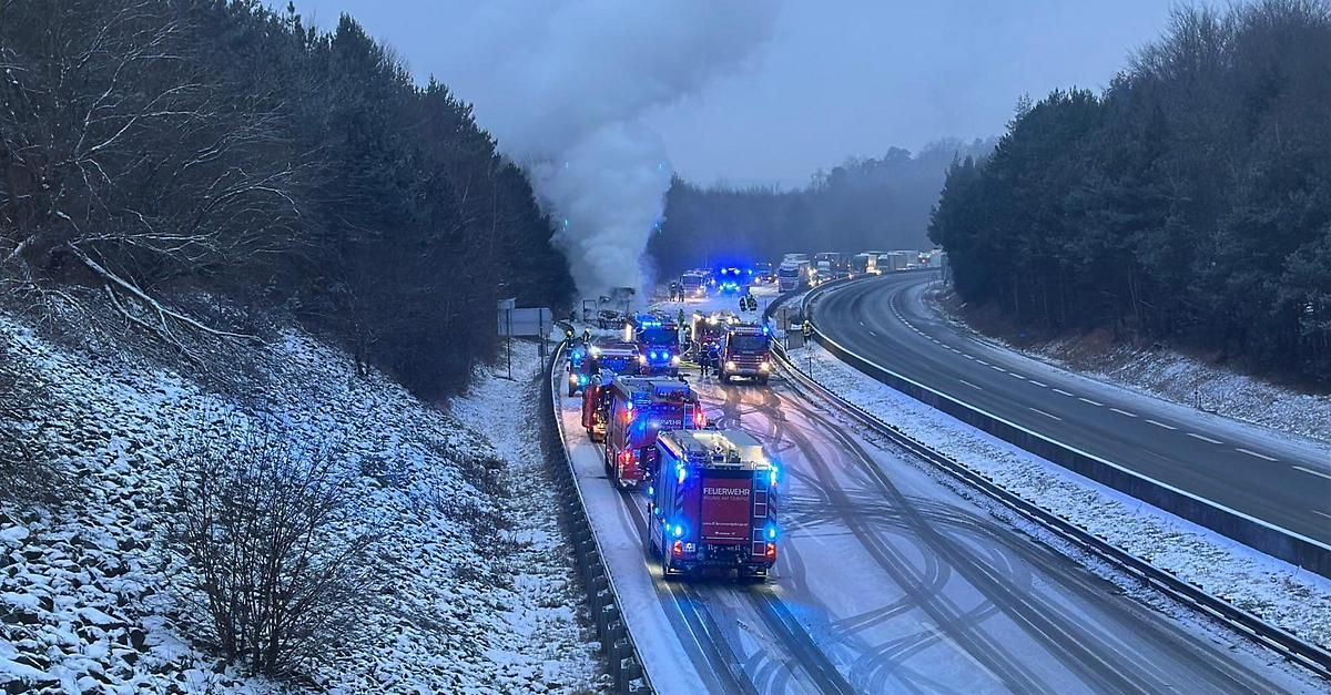 Eine verschneite Autobahn mit Rettungsfahrzeugen am Straßenrand. Rauch steigt von einem beschädigten Fahrzeug am Straßenrand auf.