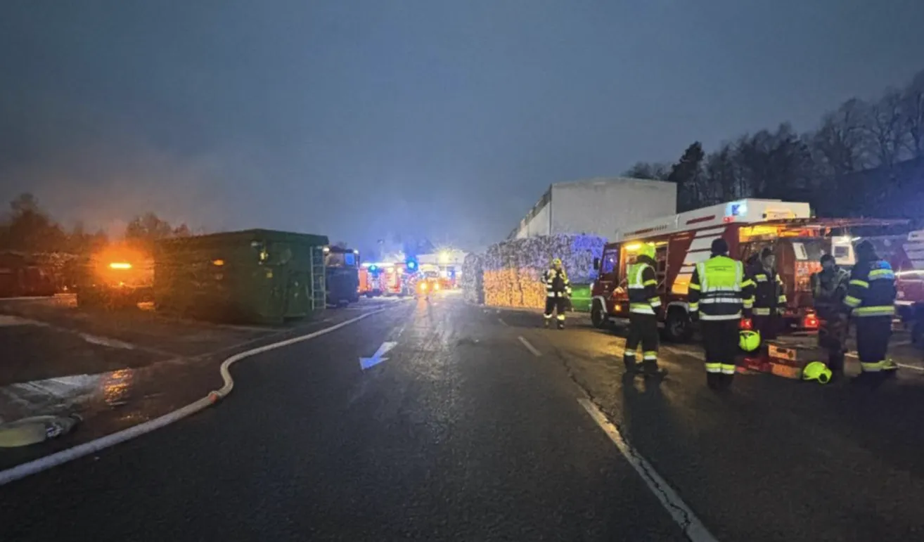 Feuerwehrleute stehen auf einer Straße, mit geparkten Einsatzfahrzeugen, einem Gebäude im Hintergrund und einem Feuerwehrwagen im Vordergrund.