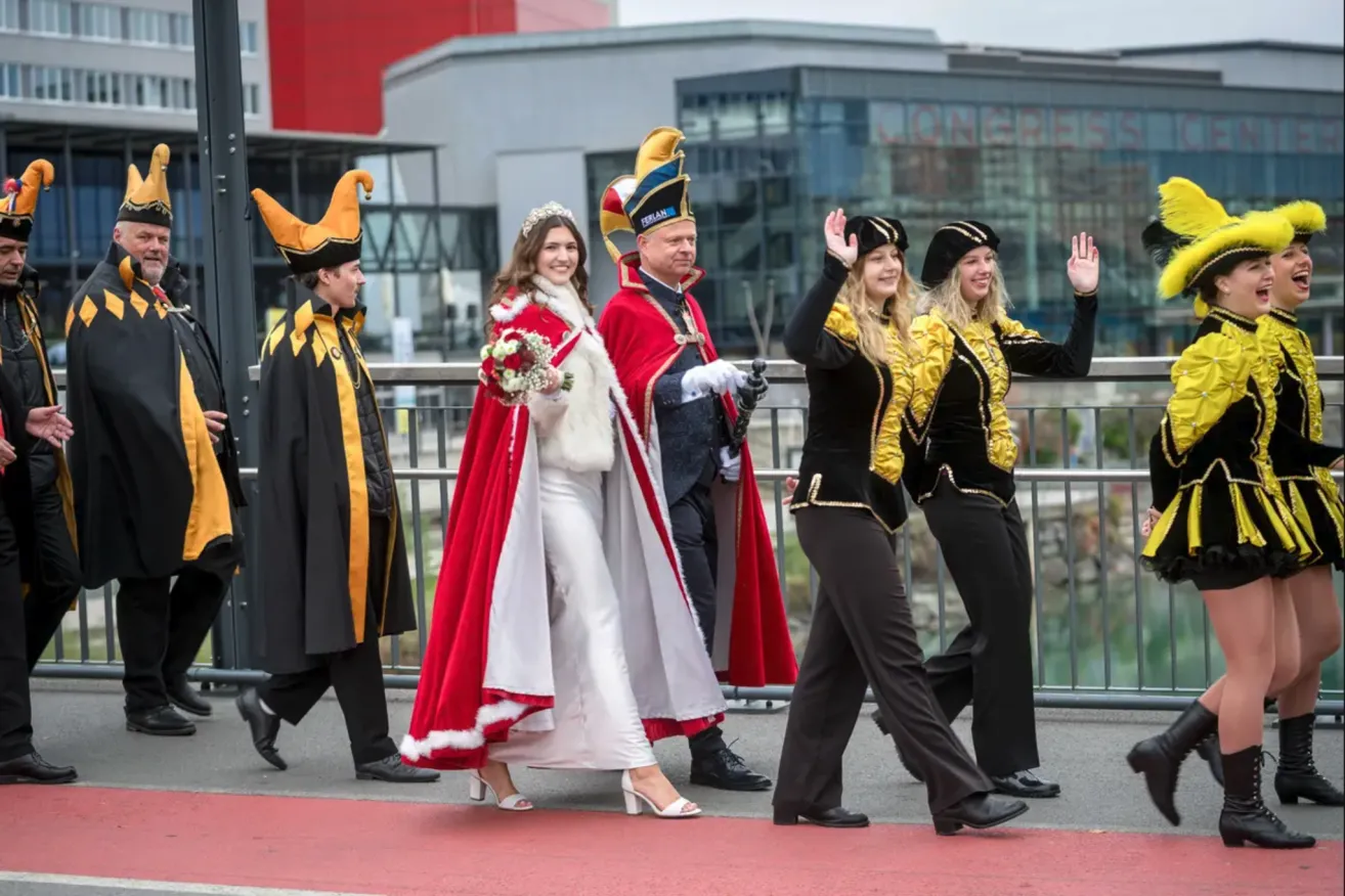 Eine Gruppe von Menschen in Kostümen, möglicherweise für einen Umzug, geht auf einem roten Teppich. Eine Frau in einem weißen Kleid hält einen Blumenstrauß. Hinter ihr winkt ein Mann in einem roten Umhang. Zwei Frauen in schwarzen und goldenen Kostümen winken. Dahinter ein Mann mit gelbem Hut. Im Hintergrund Gebäude.