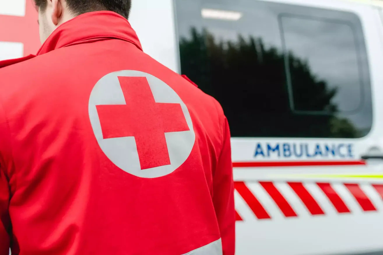 Eine Person in einem roten Ambulanzmantel mit rotem Kreuz-Symbol steht neben einem Krankenwagen.