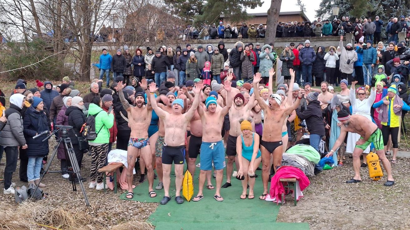 Eine Gruppe von Menschen, überwiegend Männer, steht auf einem grünen Teppich. Einige heben ihre Hände. Eine Frau in einem blauen Badeanzug steht in der Mitte. Hinter ihnen schauen Menschen zu. Einige von ihnen tragen Mützen und halten ihre Telefone.