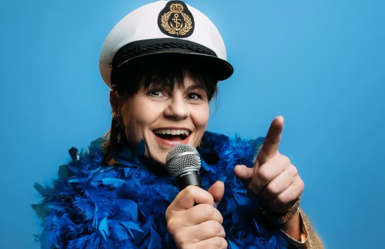 Eine Frau mit kurzen Haaren, die eine marineblaue Boa, einen Matrosenhut trägt und ein Mikrofon in der Hand hält, lächelt und zeigt mit der anderen Hand.
