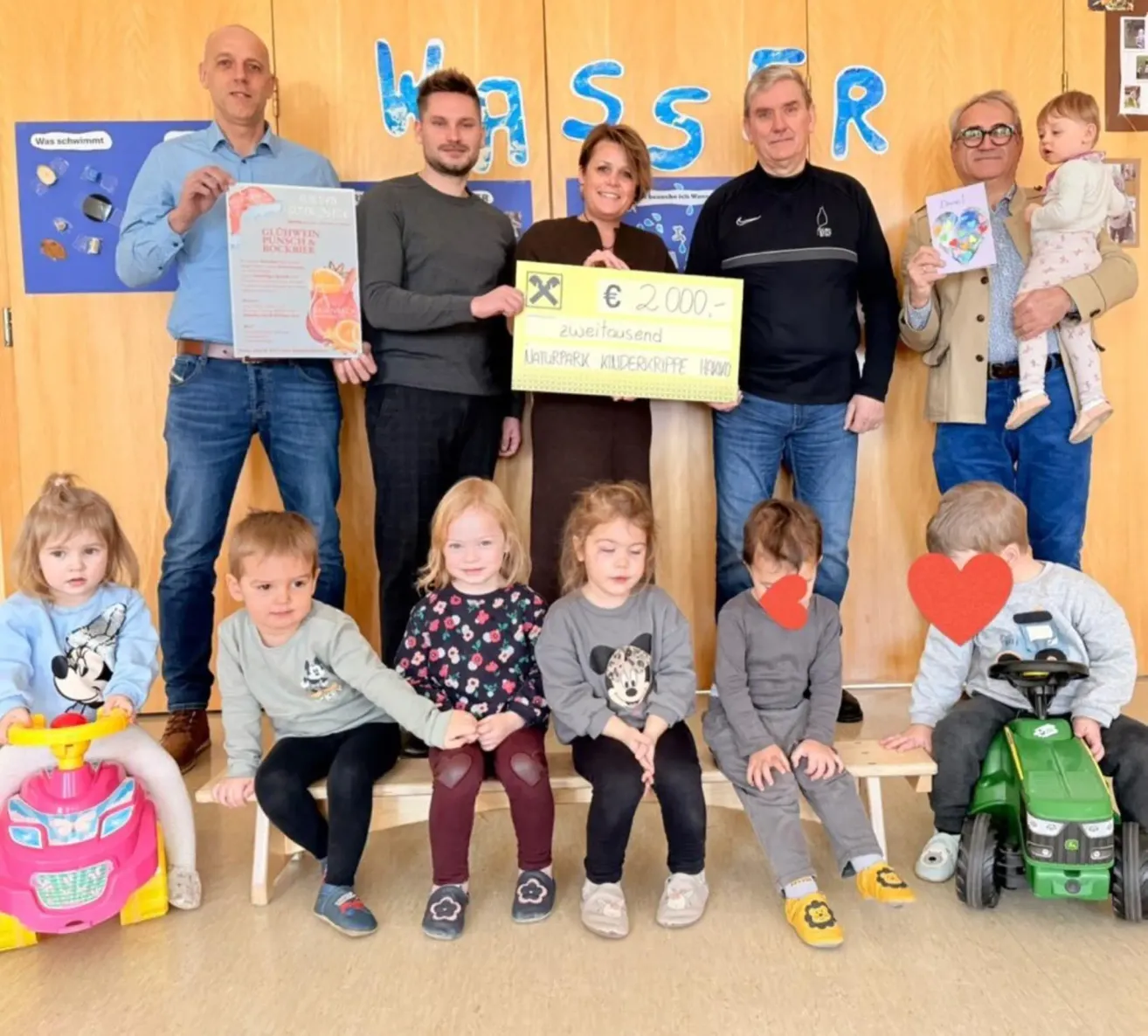 Eine Gruppe von Erwachsenen und Kindern posiert für ein Foto. Erwachsene halten Urkunden und einen Scheck. Kinder sitzen auf Bänken, und einige halten Spielzeug. Eine blaue Wand mit Text steht dahinter.