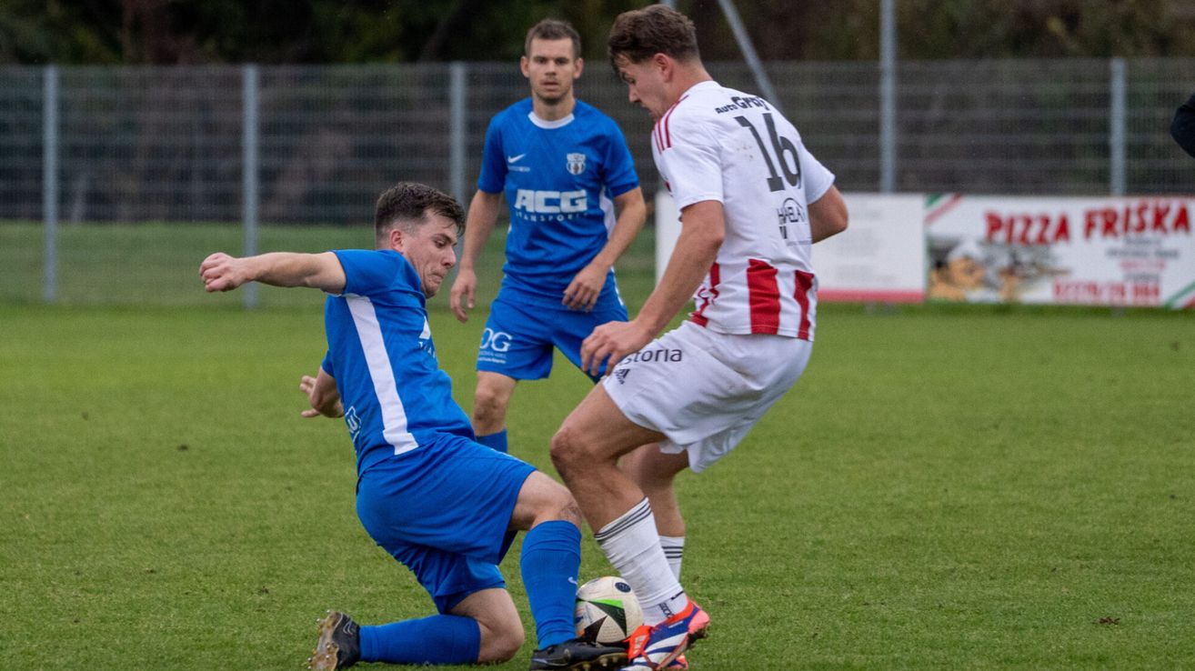 Drei Fußballspieler auf einem Rasenfeld, einer in Blau und einer in Weiß, beide tragen Trikots mit Nummern. Der Spieler in Blau liegt auf dem Boden, während der Spieler in Weiß ihn ansieht.