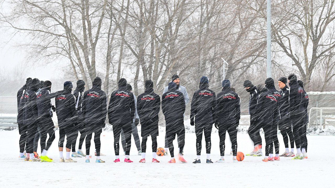Eine Gruppe von Menschen in Winterkleidung steht auf einem verschneiten Feld, trägt passende schwarze Jacken mit rotem Text und hat zwei orangefarbene Fußballbälle vor sich.