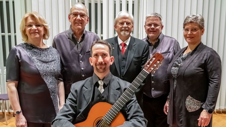 Fünf Männer stehen in einer Reihe und lächeln für ein Foto. Der Mann vor ihnen hält eine Gitarre. Die Männer hinter ihnen tragen formale Kleidung.