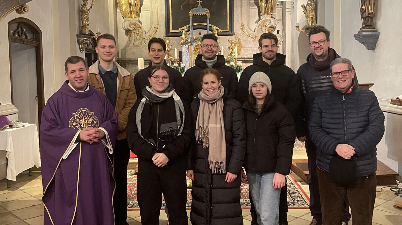 Eine Gruppe von Menschen steht in einer Kirche und lächelt für ein Foto. Sie tragen alle Winterkleidung. Der Altar ist hinter ihnen mit Kerzen und Statuen.