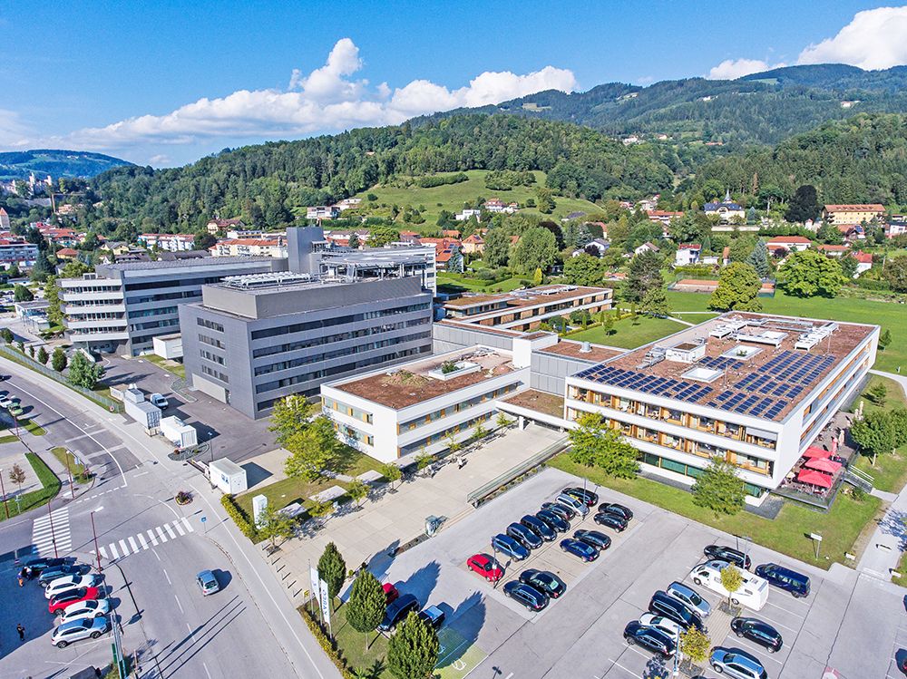 Luftaufnahme einer modernen Einrichtung mit Solarmodulen, umgeben von einer üppigen grünen Landschaft und einem geschäftigen Parkplatz mit mehreren geparkten Autos.