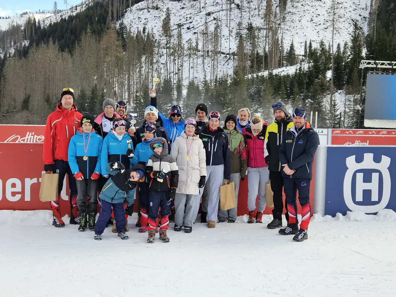 Eine Gruppe von Skifahrern in Winterkleidung steht auf einem verschneiten Hang mit einem verschneiten Berg im Hintergrund. Sie posieren für ein Foto, einige halten Trophäen und Medaillen.