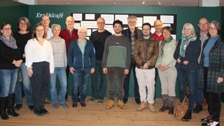 Eine Gruppe von Menschen, sowohl Männer als auch Frauen, stehen vor einer grünen Wand mit angebrachten Texten und Papieren. Sie alle lächeln und scheinen für ein Foto zu posieren.