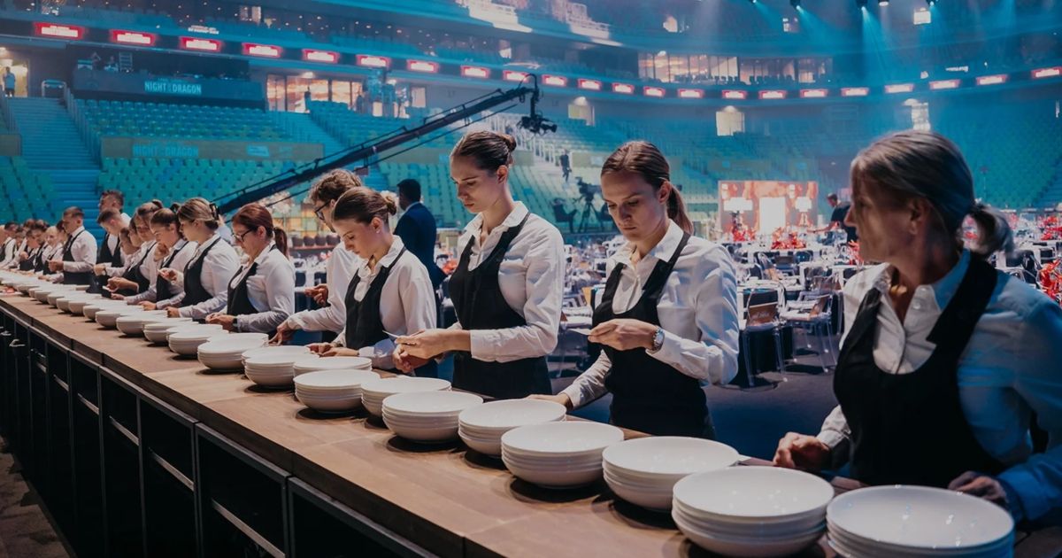 Eine Gruppe von Frauen in Schürzen stellt weiße Teller auf einem Tisch in einem leeren Stadion auf.