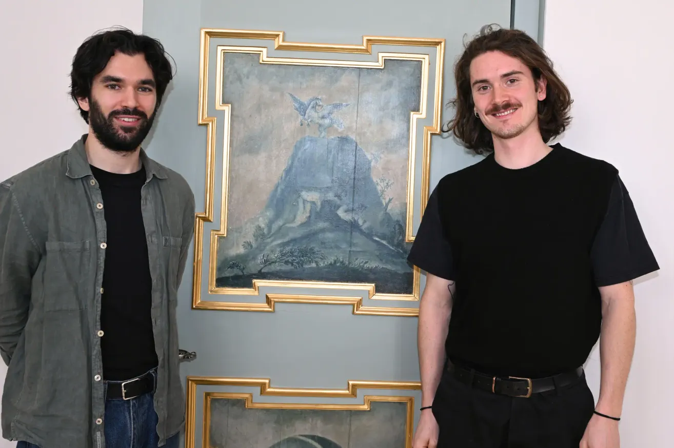 Zwei Männer stehen neben einer Wand mit zwei gerahmten Gemälden. Der Mann auf der linken Seite hat einen Bart und trägt eine Jacke. Der Mann auf der rechten Seite hat einen Schnurrbart und trägt ein schwarzes T-Shirt. Sie lächeln beide.