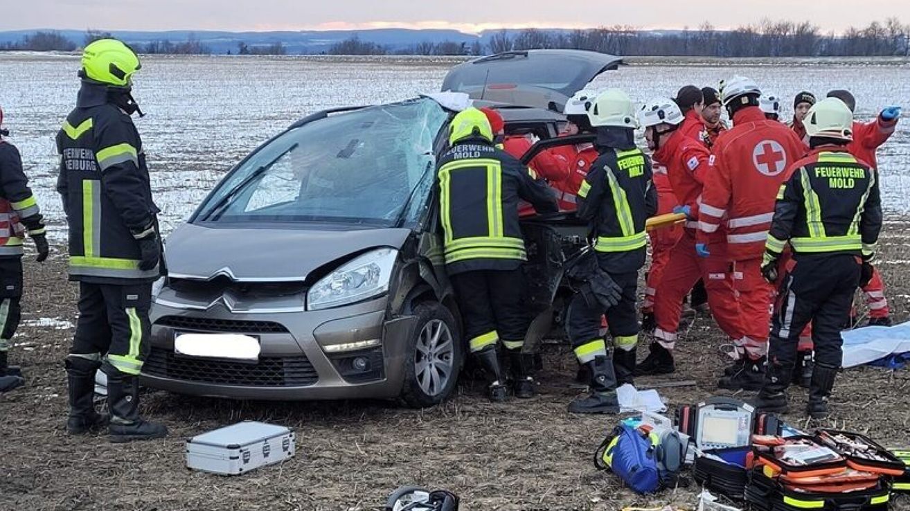 Feuerwehrleute retten einen Fahrer aus einem zerstörten Auto auf einem Feld. Sie tragen Schutzausrüstung und haben Werkzeuge in der Nähe. Das Auto ist schwer beschädigt.