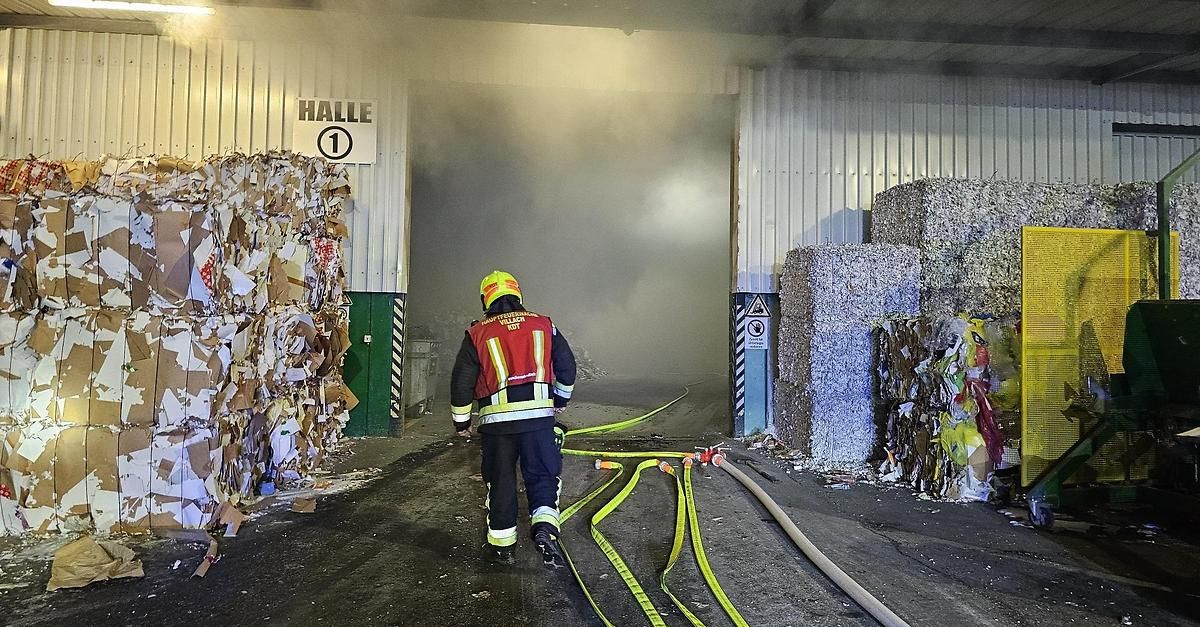 Ein Feuerwehrmann in voller Ausrüstung geht auf ein mit der Nummer 1 gekennzeichnetes Gebäude zu. Das Gebiet ist mit Rauch gefüllt, und es gibt Haufen aus Pappe und Heuballen.