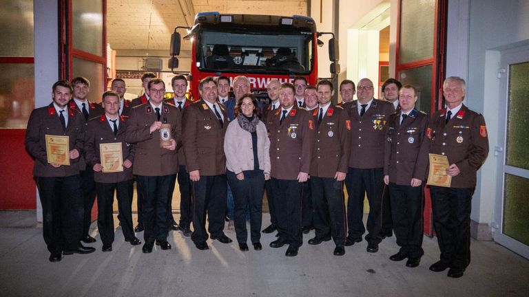 Eine Gruppe von Feuerwehrleuten posiert für ein Foto vor einem roten Feuerwehrauto, wobei ein Mann eine Flasche hält.
