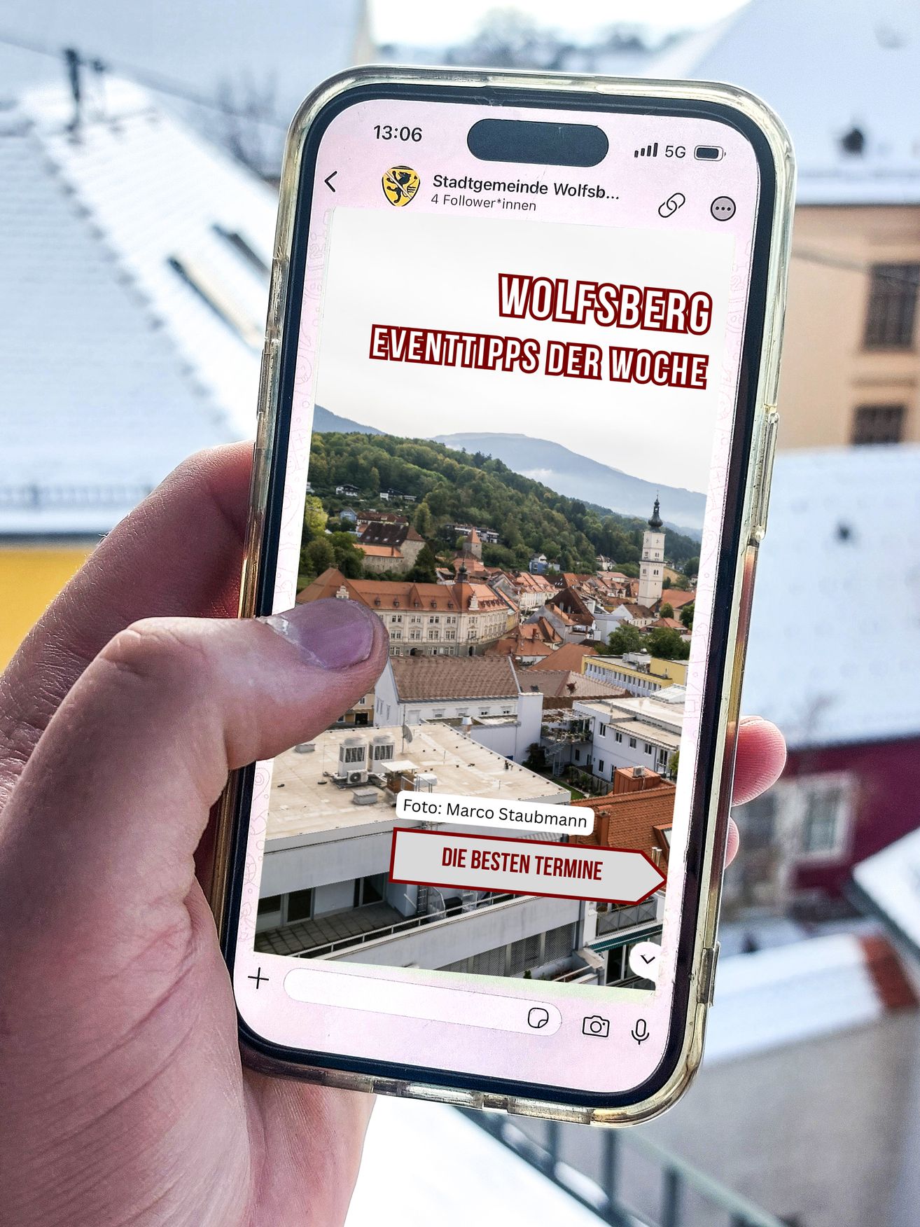 Eine Person, die ein Smartphone hält, zeigt eine App mit der Stadt Wolfsberg im Hintergrund. Der Bildschirm zeigt die Überschrift 'Wolfsberg Eventtipps der Woche' und ein Foto mit der Bildunterschrift 'Die besten Termine'.