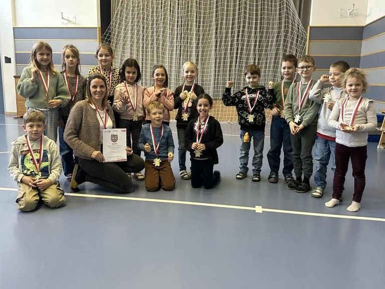 Eine Gruppe von Kindern und einer erwachsenen Frau steht mit Medaillen in einem Fitnessstudio und lächelt, mit einem weißen Vorhang und Netz hinter ihnen.