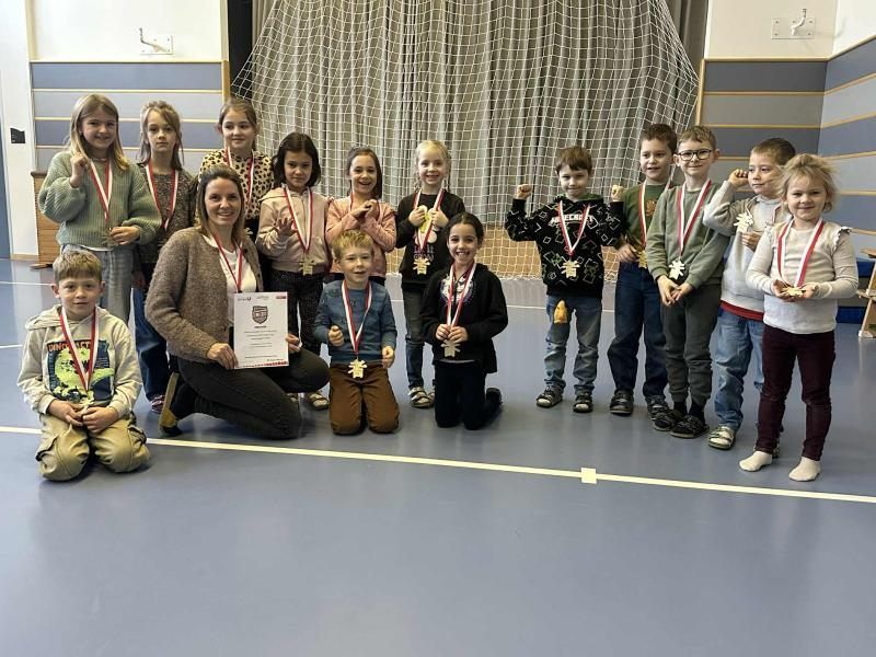 Eine Gruppe von Kindern und einer erwachsenen Frau steht mit Medaillen in einem Fitnessstudio und lächelt, mit einem weißen Vorhang und Netz hinter ihnen.