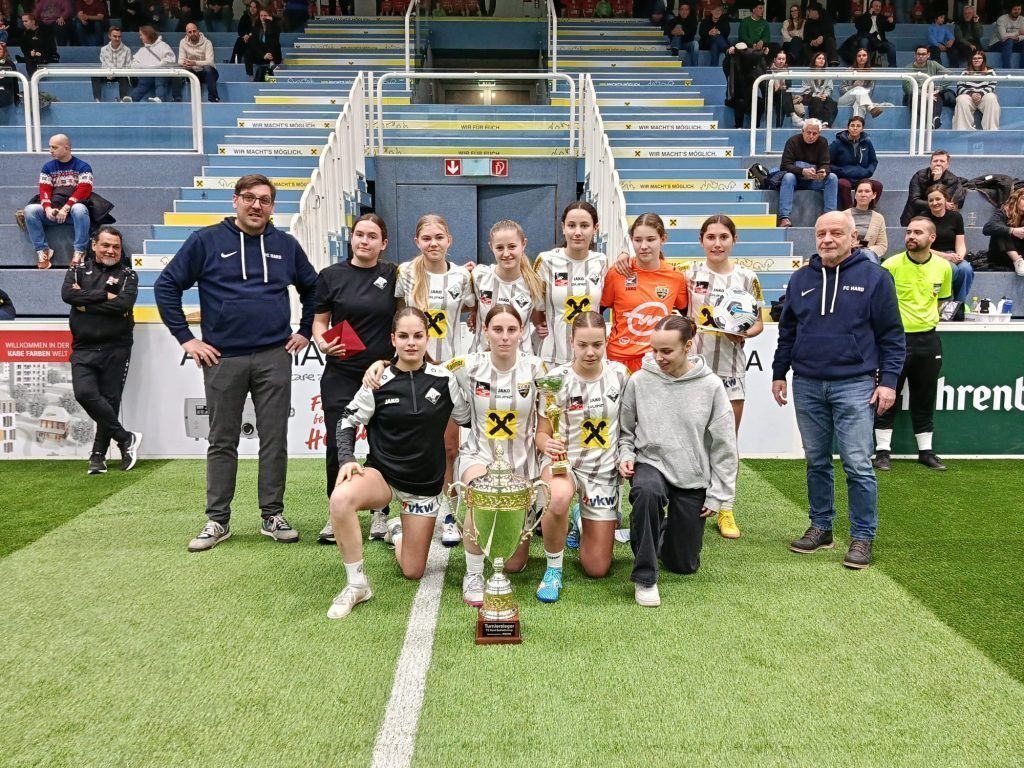 Eine Gruppe junger Frauen in Uniformen posiert mit einer Trophäe in einer überdachten Sportarena. Sie werden von zwei Männern umgeben, und im Hintergrund sitzen Menschen auf blauen Tribünen.