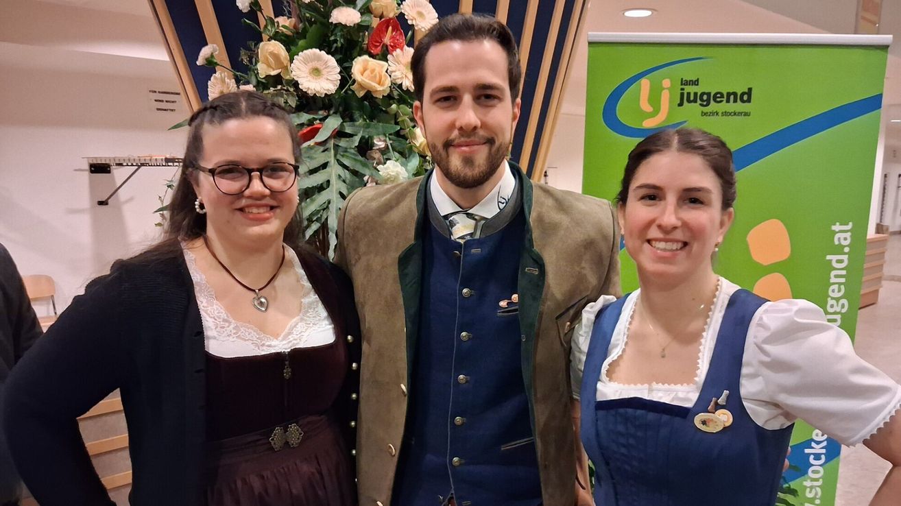 Drei Menschen lächeln in die Kamera. Ein Mann in der Mitte trägt ein traditionelles deutsches Outfit. Zwei Frauen neben ihm tragen traditionelle deutsche Kleider.