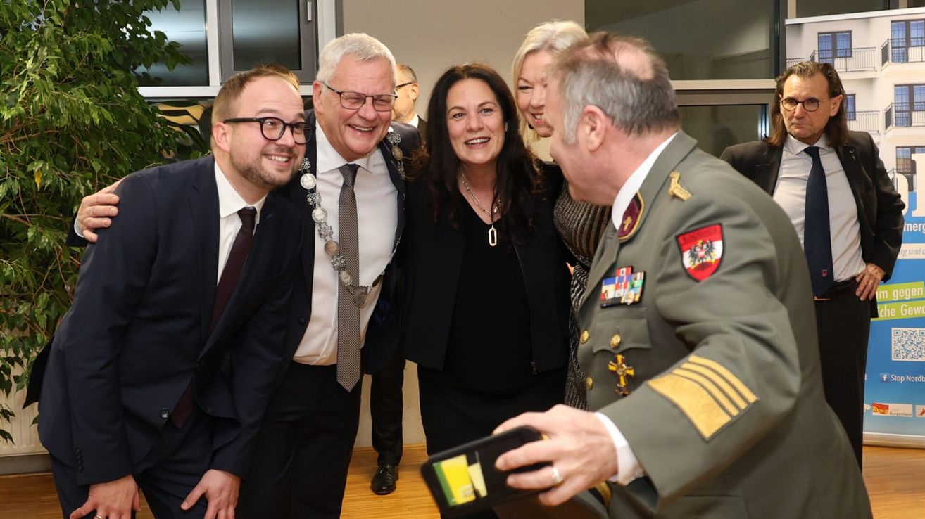 Eine Gruppe formell gekleideter Personen, darunter ein Mann in Uniform, ein Mann mit Kette und eine Frau mit Halskette, lächeln und scheinen in einem Raum zu plaudern.
