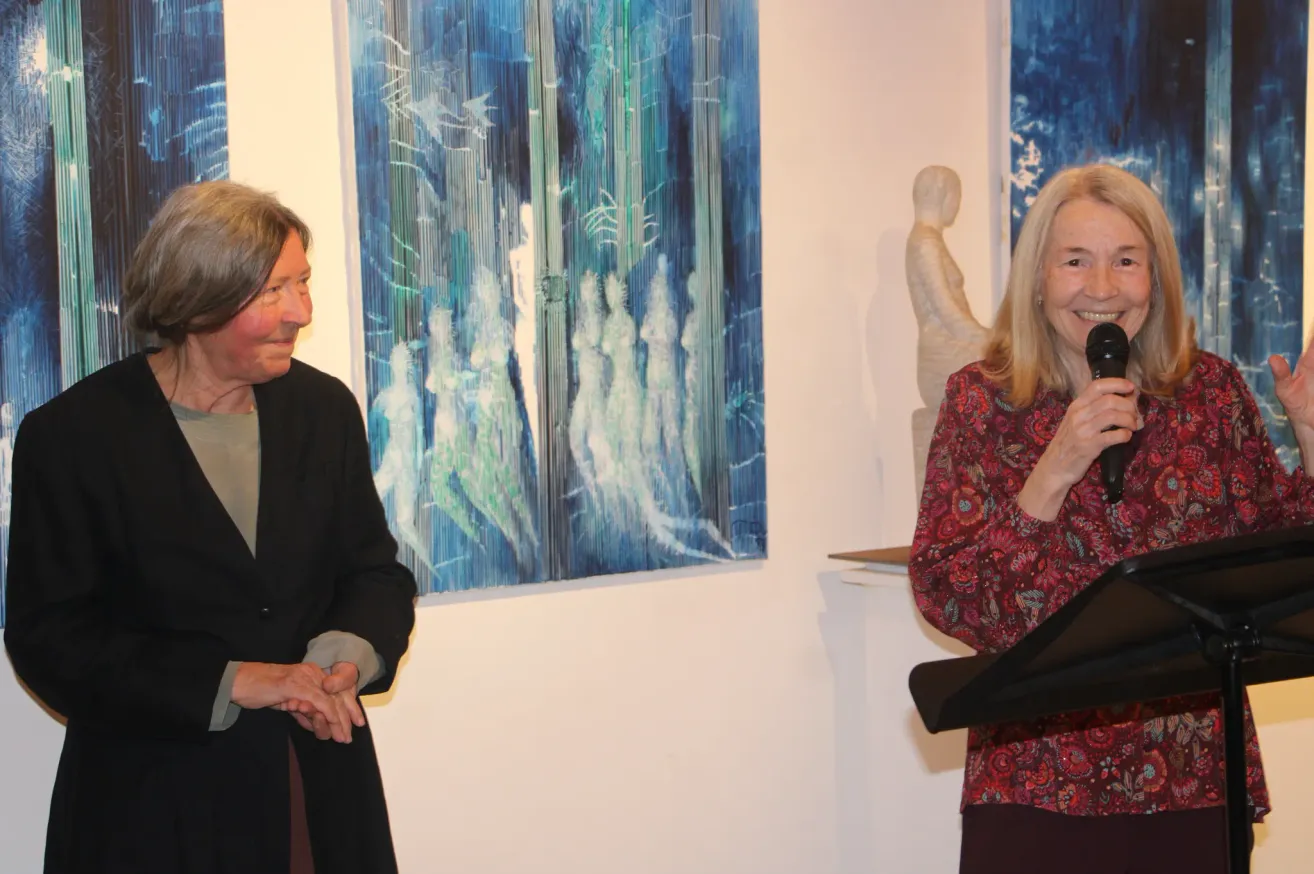 Zwei Frauen befinden sich in einer Kunstgalerie. Eine Frau hält ein Mikrofon und spricht. Hinter ihr ist ein blaues Gemälde.