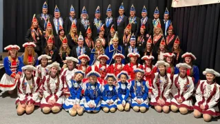 Eine Gruppe von Menschen in traditionellen Kostümen und Hüten posiert für ein Foto. Einige sitzen auf dem Boden, während andere stehen. Sie tragen alle Medaillen um den Hals.