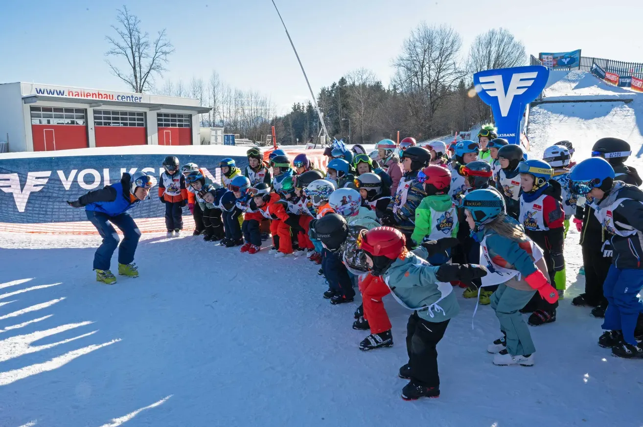 Eine Gruppe von Kindern in Skiausrüstung steht in einer Reihe auf einem verschneiten Feld, möglicherweise an einem Ski-Event teilnehmend.