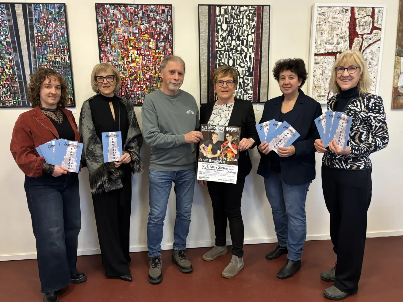 Eine Gruppe von sechs Personen steht in einer Kunstgalerie. Sie halten ein Buch und einen Flyer. Sie tragen alle Brillen und Lederschuhe. Hinter ihnen hängen drei Gemälde. Die mittlere Person hält ein Plakat mit den Worten 'Aus Wertham Himmel'. Der Boden ist aus Holz.