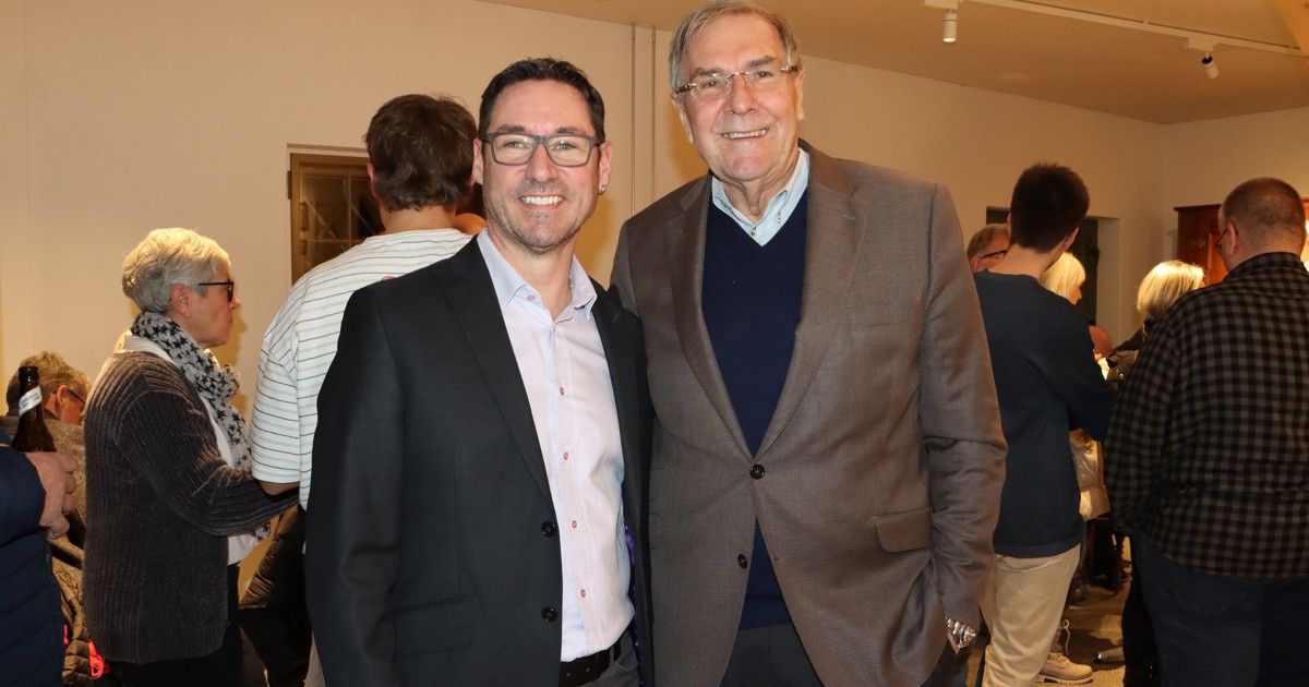 Zwei Männer in Anzügen lächeln für ein Foto, einer trägt eine Brille und der andere einen hellen Blazer. Hinter ihnen schauen ein Mann in einem gestreiften Hemd und ein anderer in einem schwarzen Hemd zu.