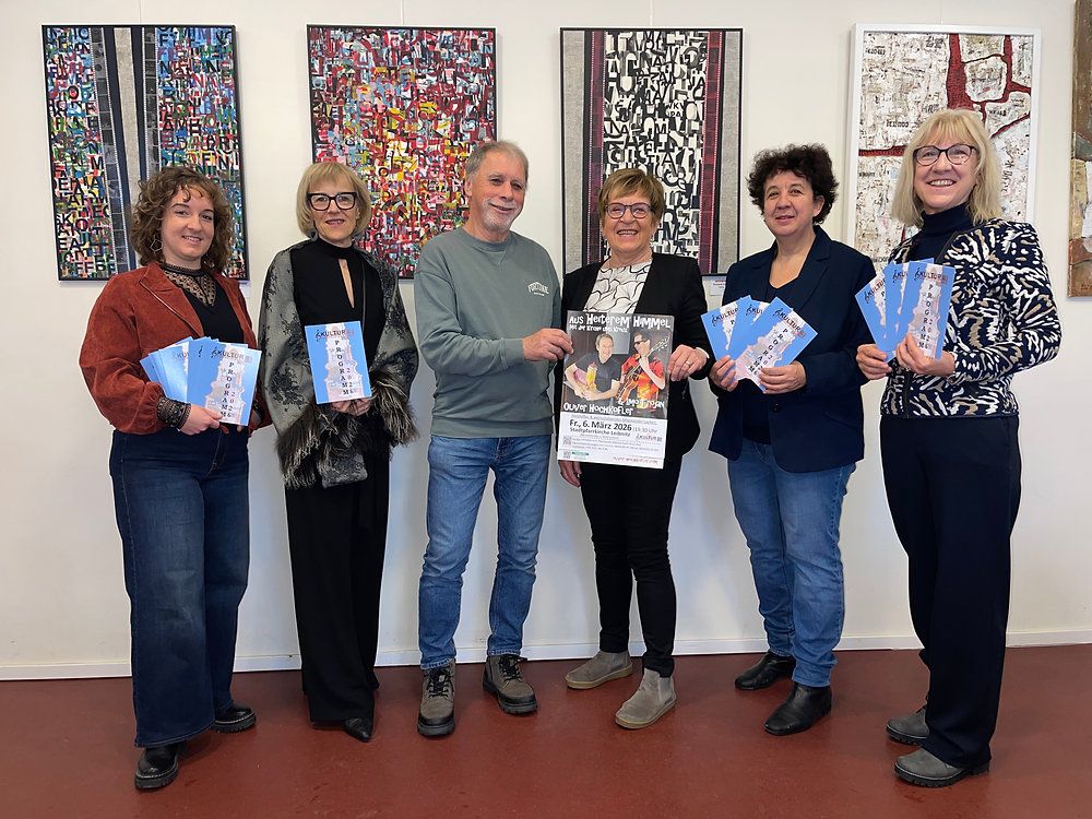 Sechs Personen stehen zusammen und halten Eintrittskarten und Broschüren. Eine Frau in der Mitte hält eine Broschüre mit einem Bild eines Mannes und Text über eine Veranstaltung am 6. März 2026.