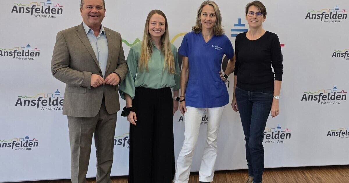 Vier Personen stehen vor einem weißen Hintergrund, zwei Frauen in lässiger Kleidung und zwei Männer, einer in einem Anzug und der andere in einer medizinischen Uniform. Sie alle lächeln und scheinen für ein Foto zu posieren.