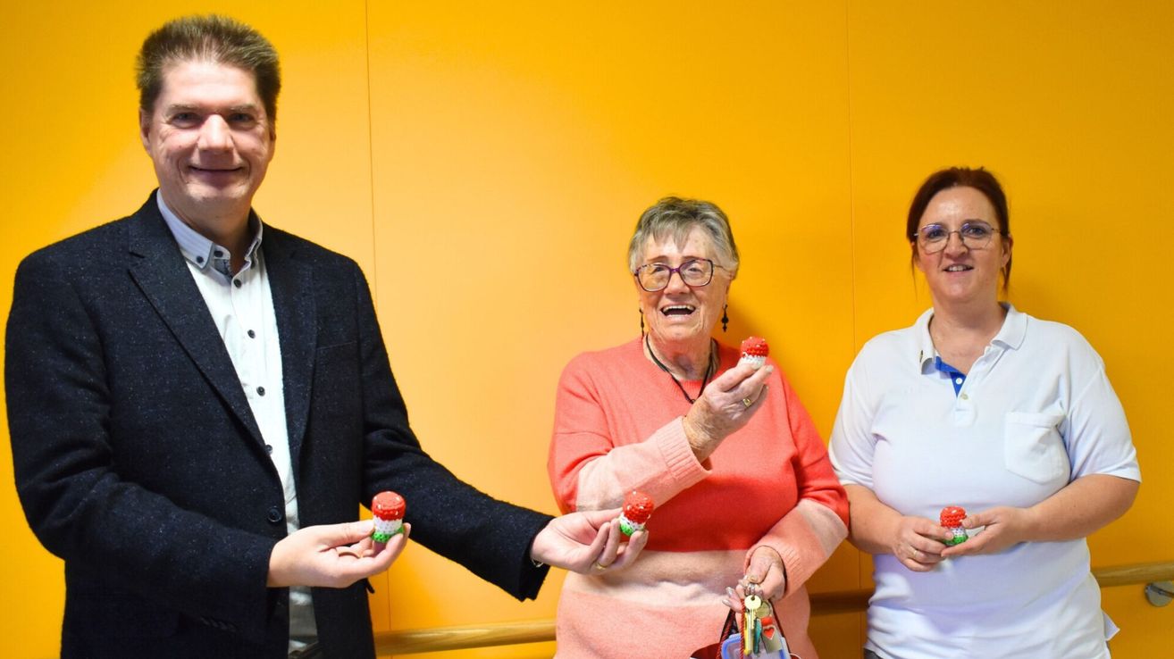 Drei Personen stehen vor einer gelben Wand. Die Frau in der Mitte, mit Brille, lächelt und hält kleine gestrickte Objekte. Der Mann links hält ein ähnliches Objekt. Die Frau rechts trägt ein weißes Hemd.