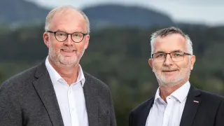 Zwei Männer, beide mit Brille, stehen nebeneinander vor einem verschwommenen Hintergrund. Beide tragen weiße Hemden, aber einer trägt einen grauen Blazer.
