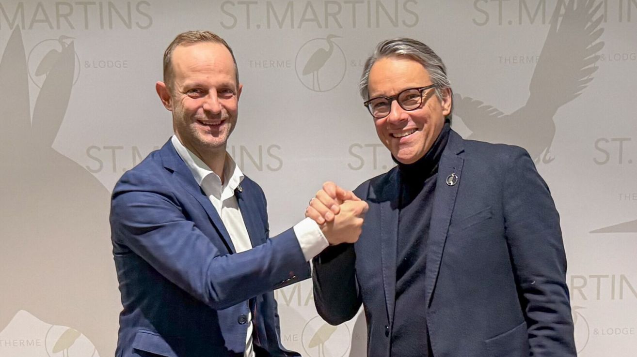 Zwei Männer in formeller Kleidung, lächelnd und Hände schüttelnd vor einer Wand mit dem Logo und Text von St. Martins Therme.