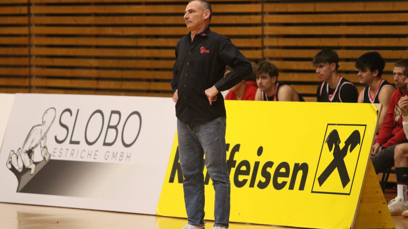 Ein Mann steht auf einem Basketballplatz mit den Händen auf dem Rücken. Hinter ihm befinden sich Holztribünen mit Zuschauern. Ein gelber Banner mit schwarzem Text ist auf dem Platz.