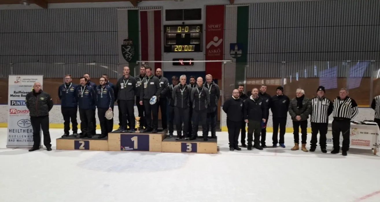 Eine Gruppe von Männern in schwarzen Jacken steht auf einem Podium in einer Eishalle, hält Medaillen und Trophäen. Hinter ihnen zeigt eine Anzeigetafel einen Punktestand von 0-0, mit einem Timer bei 20:00. Die Halle hat Glasbarrieren, und Flaggen und Banner hängen an den Wänden.
