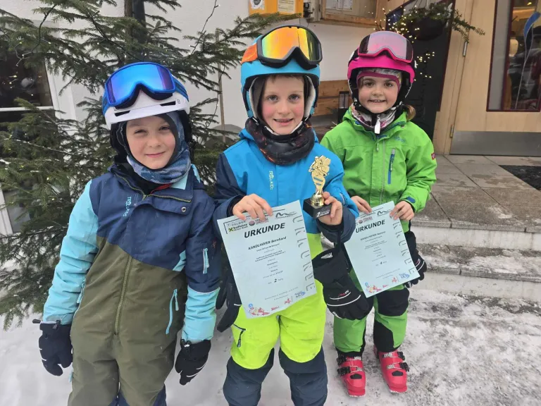 Drei Kinder in Skiausrüstung stehen im Schnee. Sie halten Urkunden und eine Trophäe in den Händen. Im Hintergrund ist ein Gebäude mit einer Weihnachtsbaum und Dekorationen zu sehen.