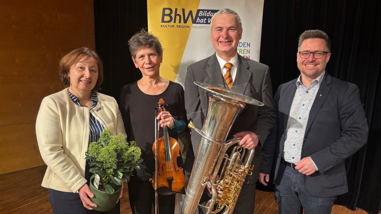 Vier Menschen stehen vor einem Banner, zwei halten Musikinstrumente, einer lächelt und ein Mann im Anzug lächelt in die Kamera.