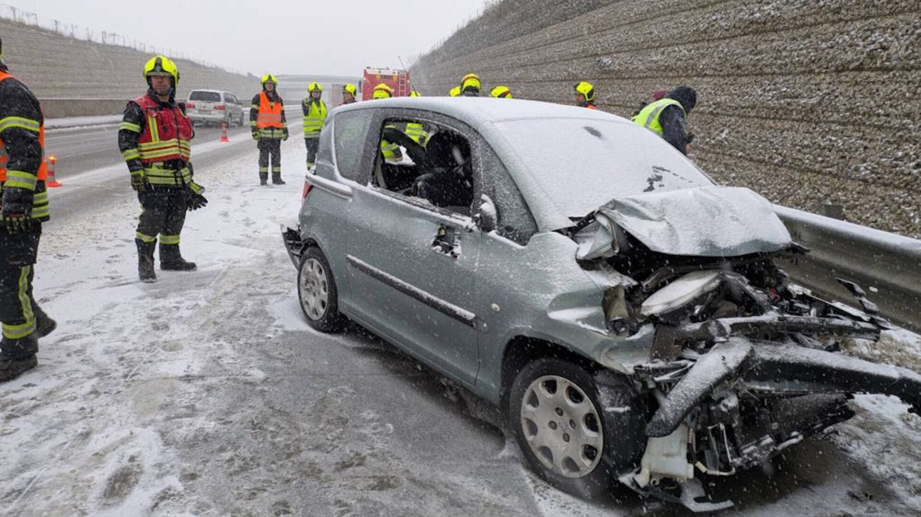 Rettungskräfte stehen um ein demoliertes Auto auf einer verschneiten Straße, mit einem Feuerwehrwagen im Hintergrund.