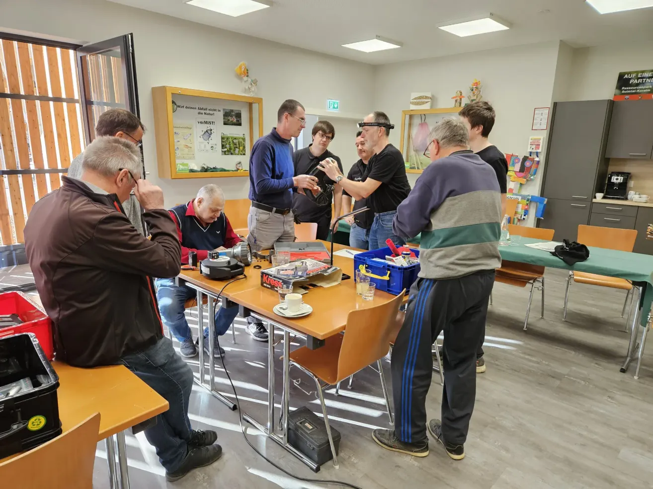 Eine Gruppe von Männern versammelt sich um einen Tisch, möglicherweise für einen Workshop. Ein Mann hält ein Werkzeug, während ein anderer in der Nähe steht. Der Raum hat Stühle, Tische und ein Glasfenster.