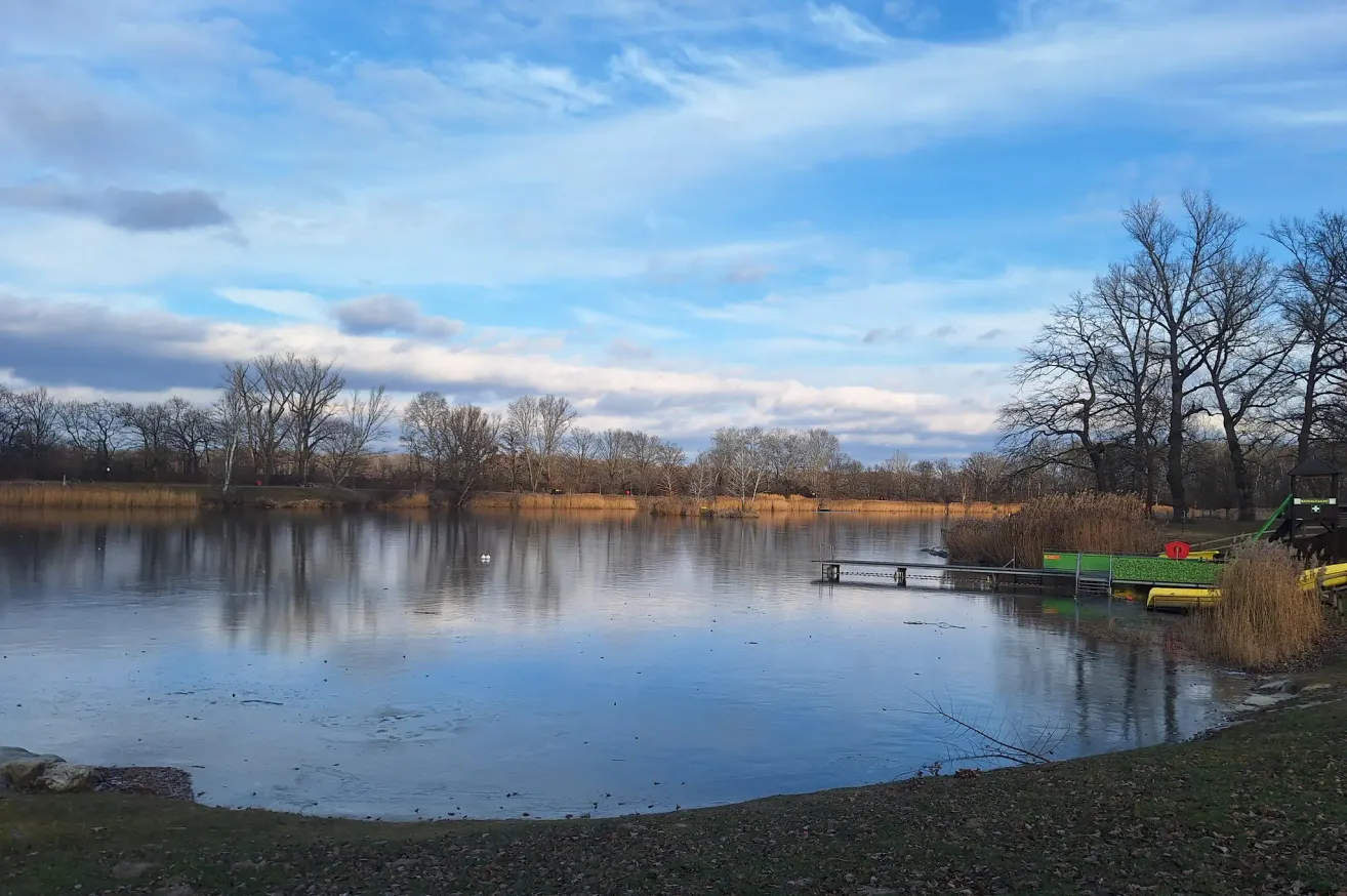 Ein ruhiger See mit klarem blauem Himmel und verstreuten Wolken. Der See ist von kahlen Bäumen umgeben, und rechts befindet sich ein kleiner Steg.