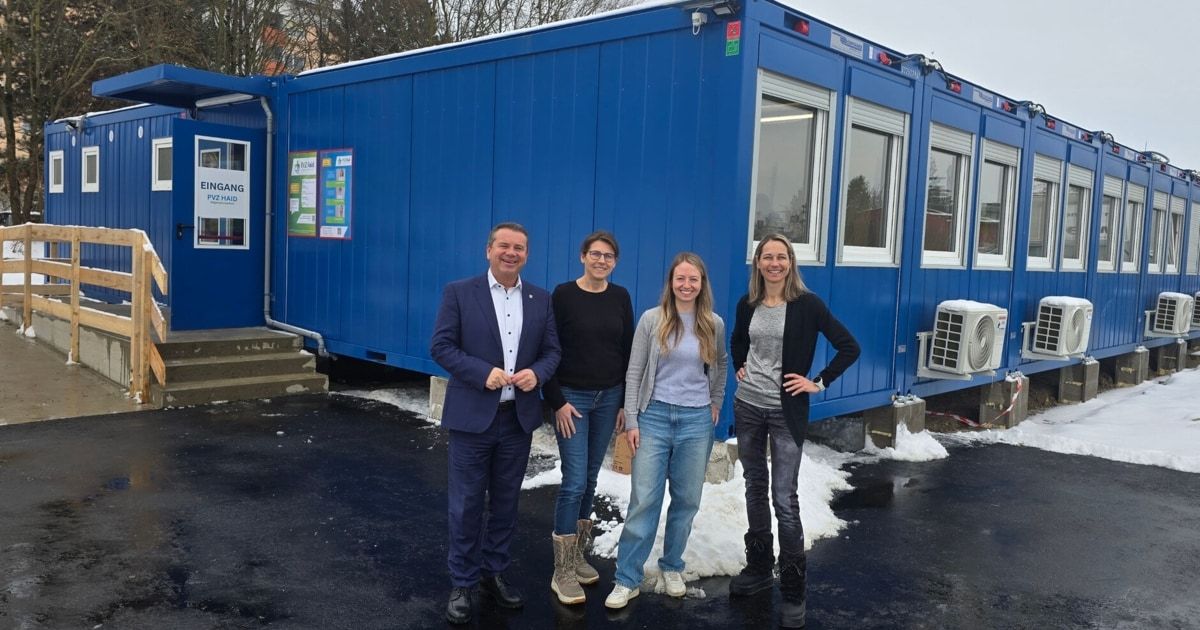 Eine Gruppe von vier Personen steht vor einem blauen Gebäude und lächelt für ein Foto. Das Gebäude hat Glasfenster und ein Schild mit der Aufschrift 'Lang Aid'. Sie sind in Winterkleidung gekleidet.