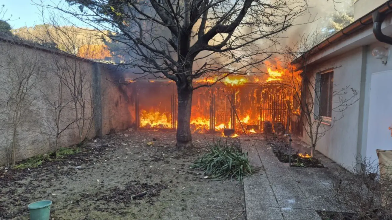 Ein Feuer verzehrt ein Haus, Flammen lodern aus Dach und Fenstern, umgeben von einer kargen Landschaft mit einem Baum und einer Steinmauer.