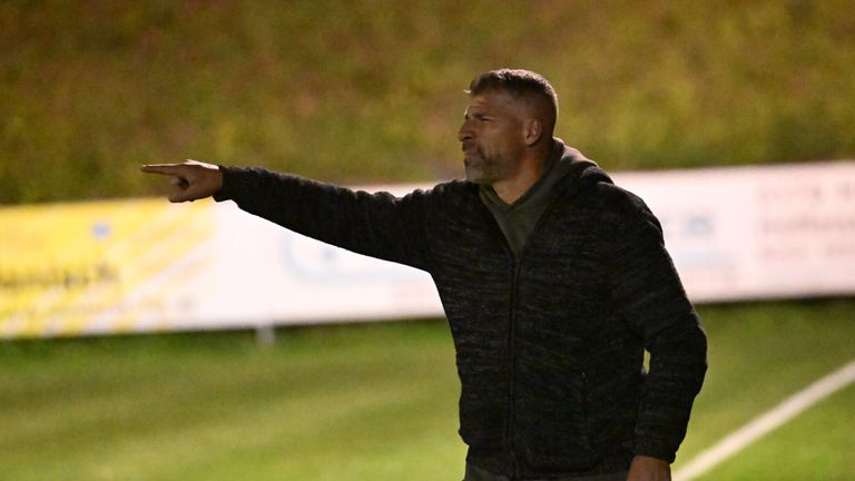 Ein Mann in einer schwarzen Kapuzenjacke zeigt auf einem Fußballfeld in der Dämmerung.