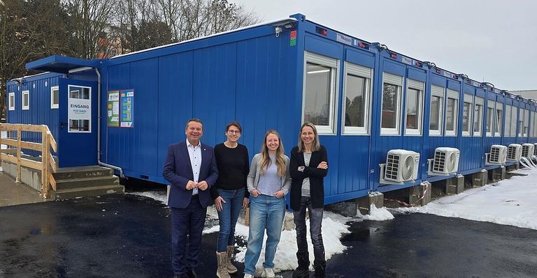 Vier Personen, zwei Männer und zwei Frauen, stehen lächelnd vor einem blauen Container für ein Foto.