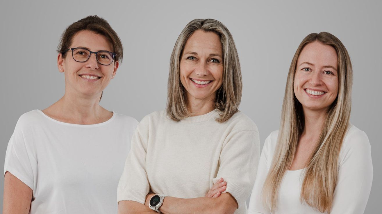 Drei Frauen stehen nebeneinander und lächeln in die Kamera, eine mit einer Brille und einem weißen T-Shirt, eine andere mit einem weißen Pullover und einer Uhr und die dritte mit einem weißen Oberteil und langen blonden Haaren.