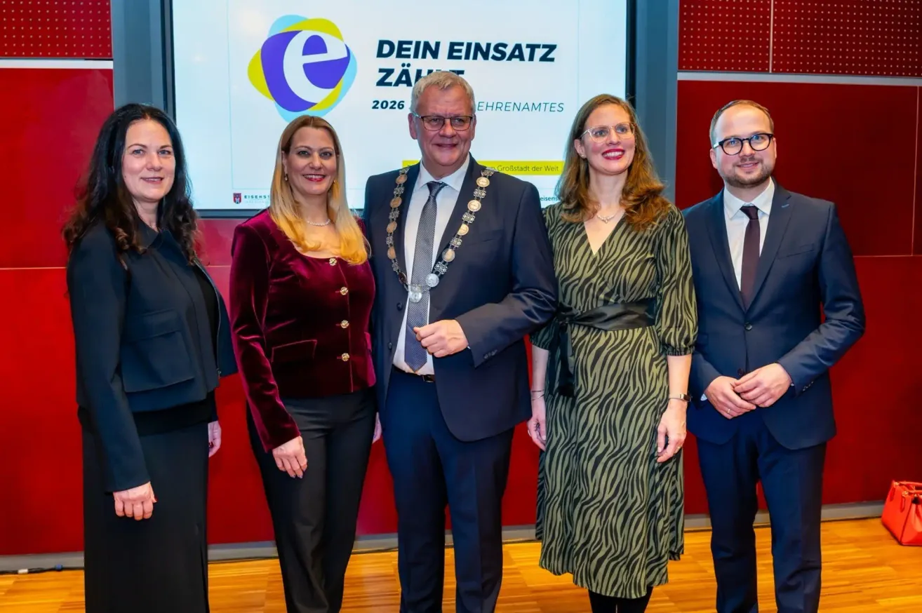 Fünf Personen stehen vor einer Leinwand mit der Aufschrift 'Dein Einsatz 2026'. Der Mann in der Mitte trägt eine Medaille und eine Brille. Die Frauen auf beiden Seiten lächeln. Hinter ihnen ist eine rote Wand zu sehen.