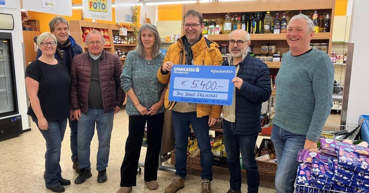 Eine Gruppe von Menschen steht in einem Lebensmittelgeschäft. Ein Mann hält einen blauen Scheck über 5.400 Euro und lächelt. Dahinter hält ein weiterer Mann ein Poster mit einem Hashtag.